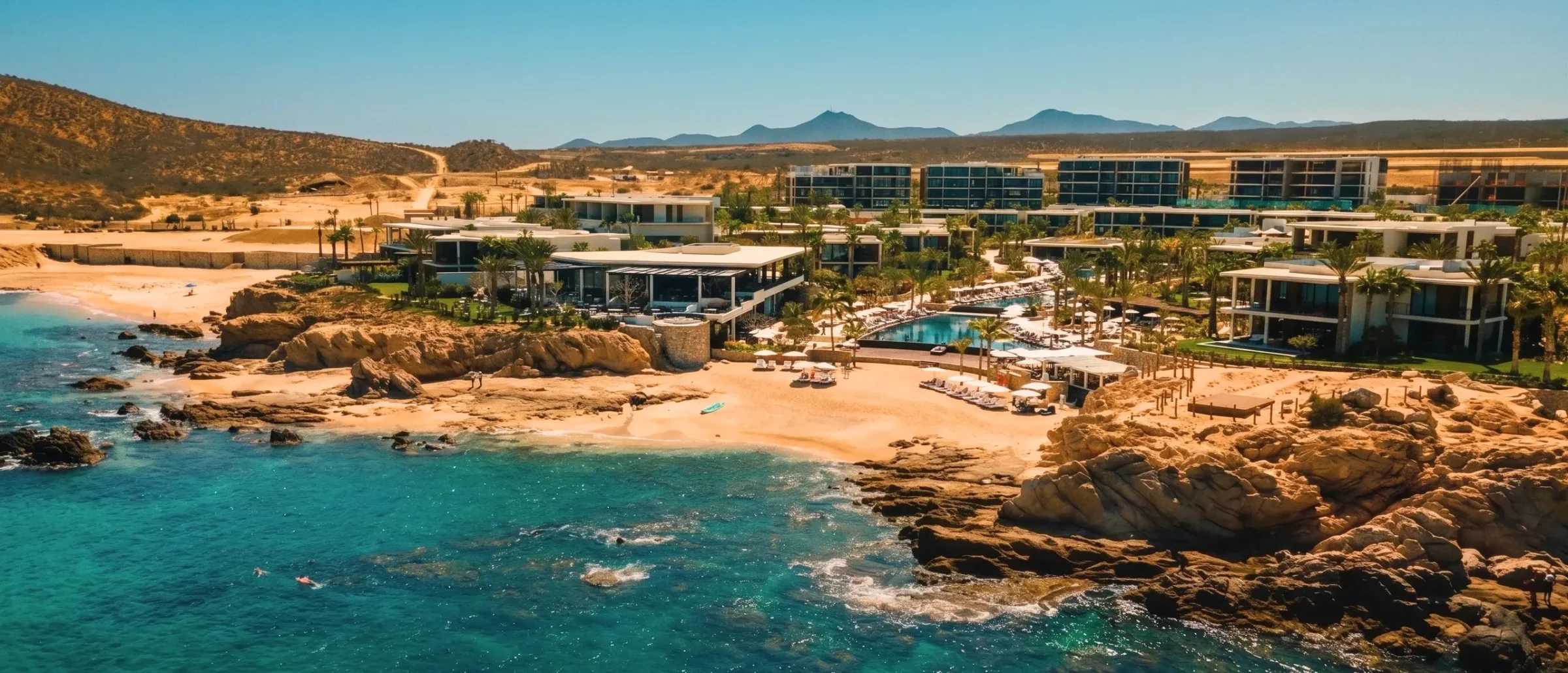 Aerial view of modern beachfront residences with a pool and palms above a rocky cove with turquoise water