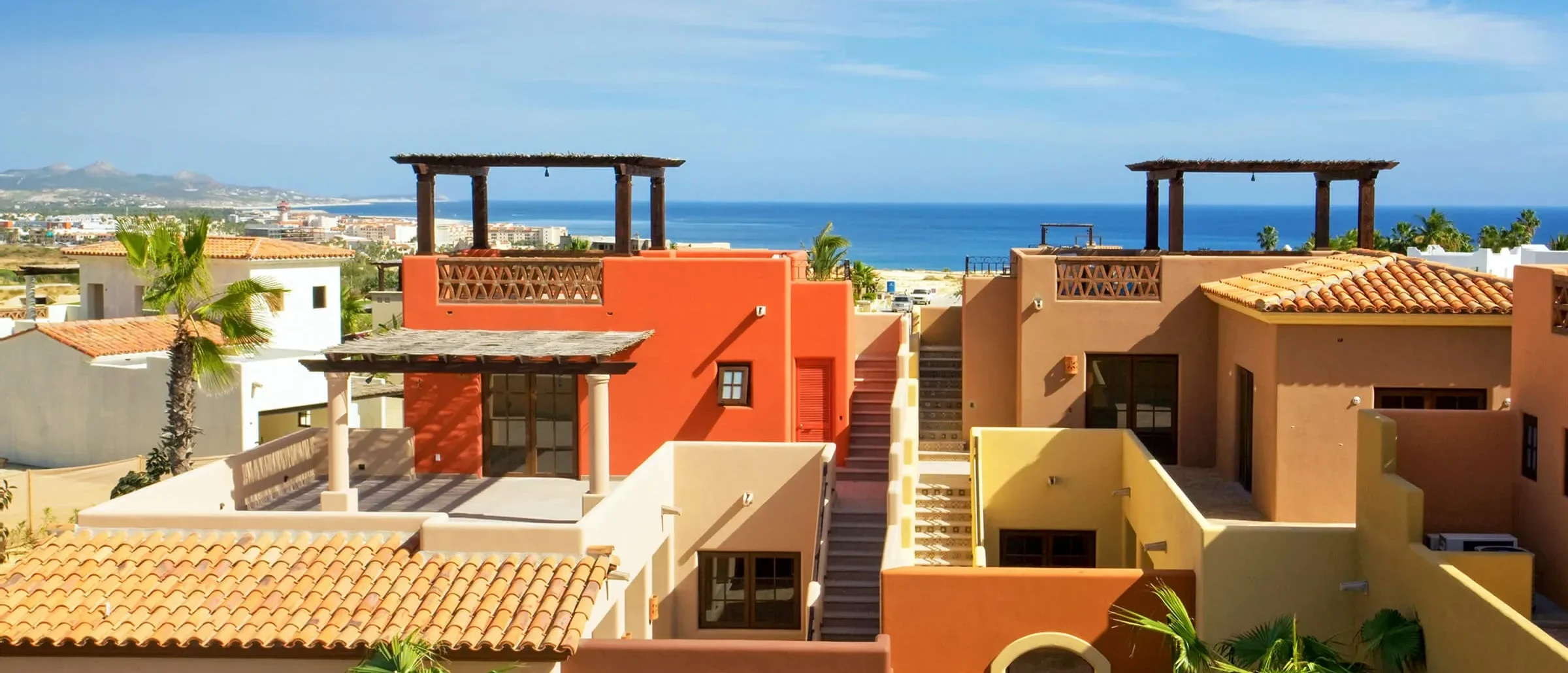 Colorful hacienda-style homes with tile roofs and rooftop pergolas, looking out to the ocean and coastline