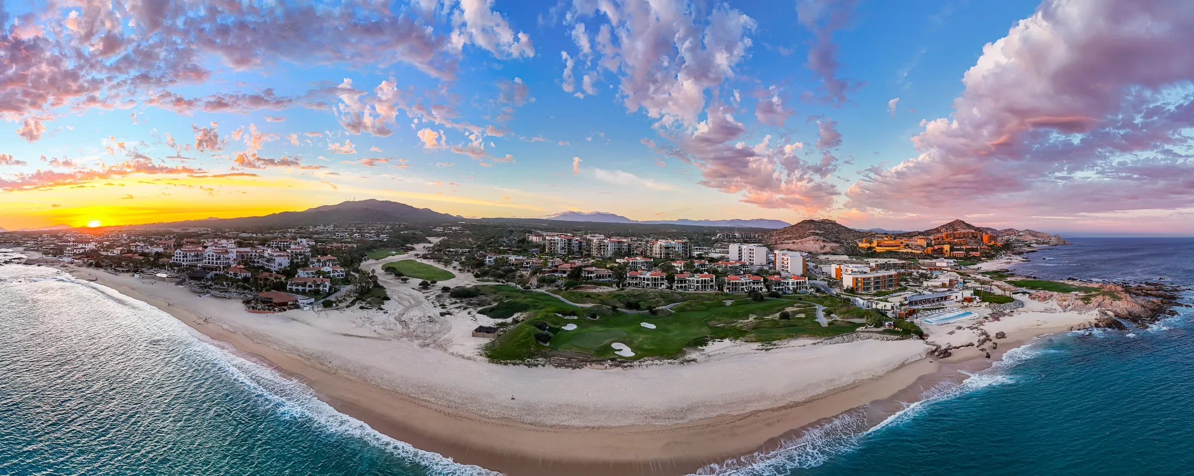 Panoramic aerial at sunset of a curved beach, oceanfront golf course and resort buildings, mountains behind