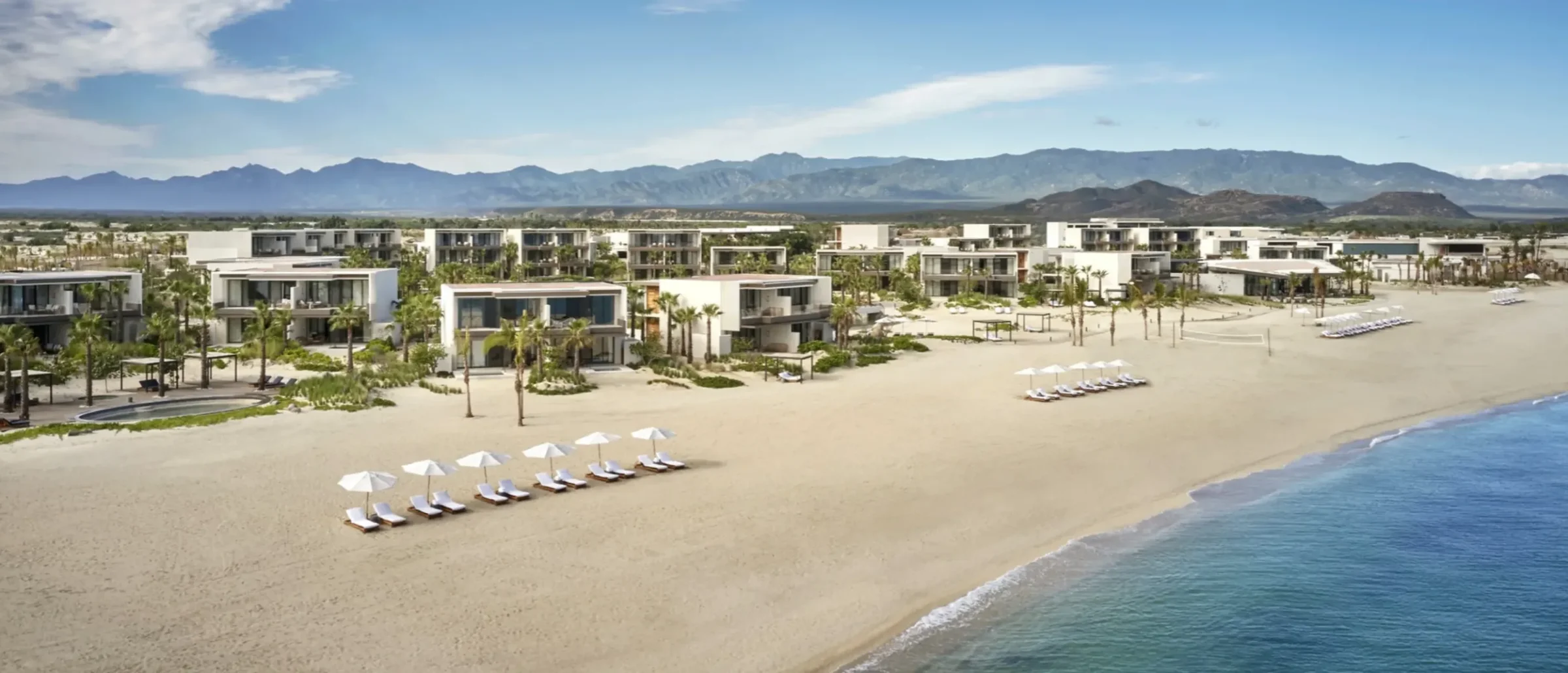 Aerial view of a beachfront resort with white umbrellas on the sand, modern villas and a mountain backdrop
