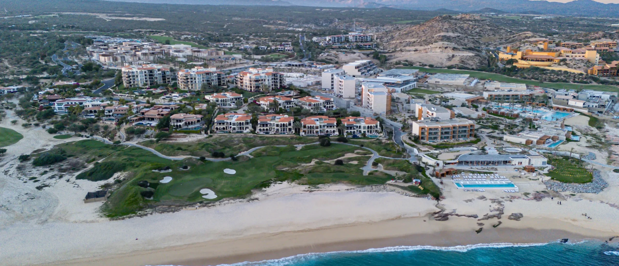 Aerial view of a beachfront golf hole, red-roofed villas, condos and pools with desert mountains behind