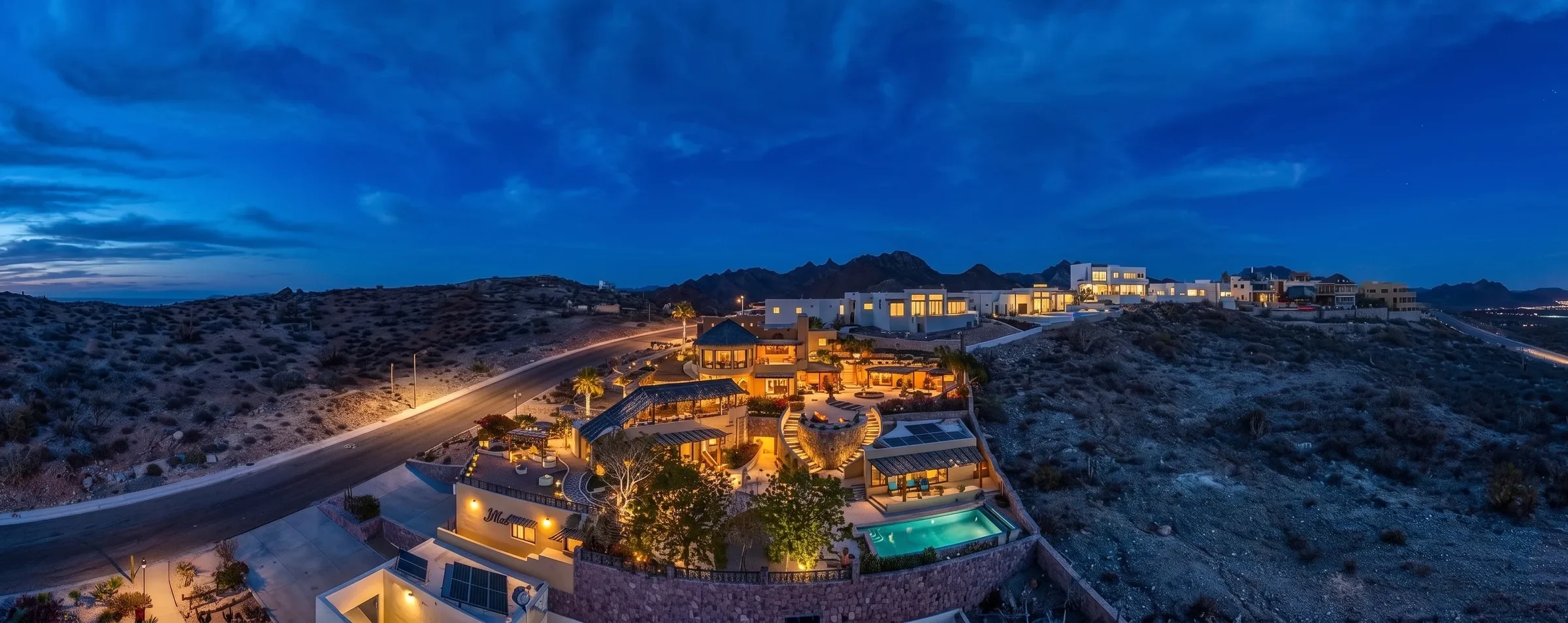 Aerial dusk panorama of a lit hillside villa with a pool, modern homes on a desert ridge behind