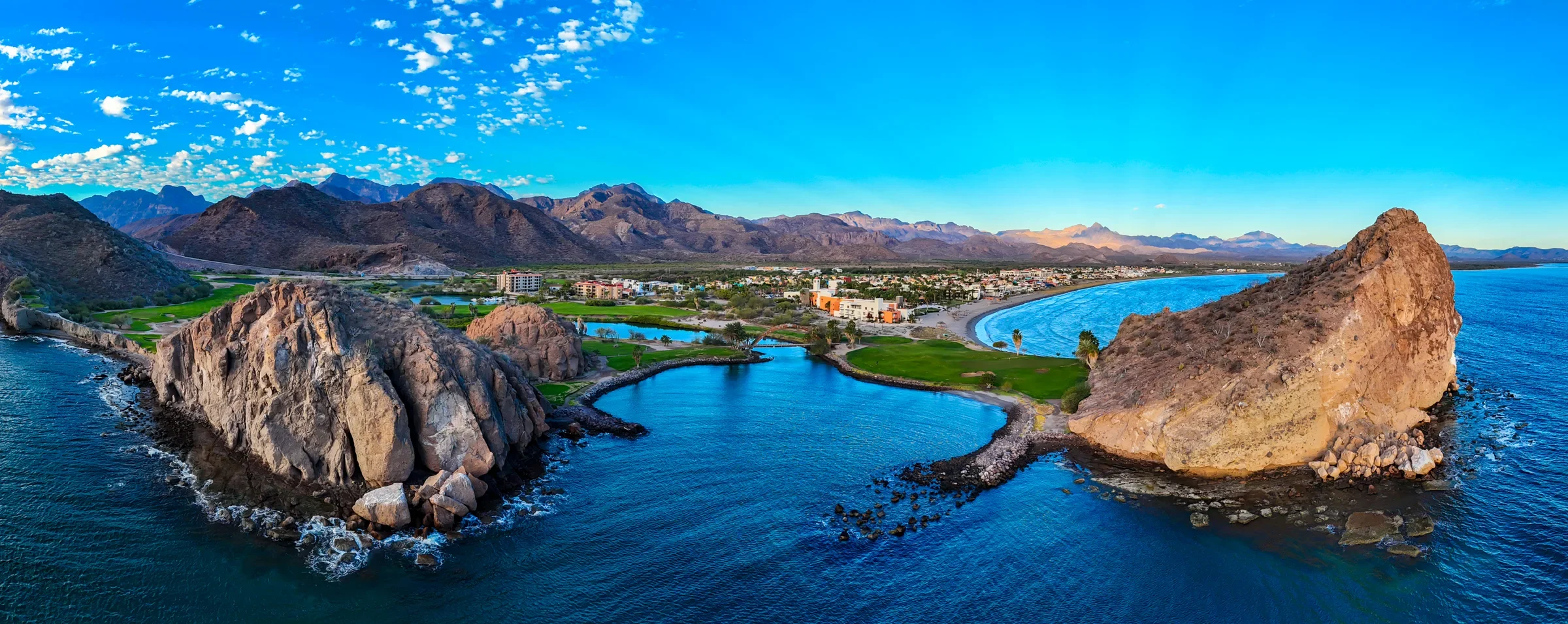 Aerial panorama of a rocky cove, golf course and low-rise coastal town below rugged mountains near Loreto