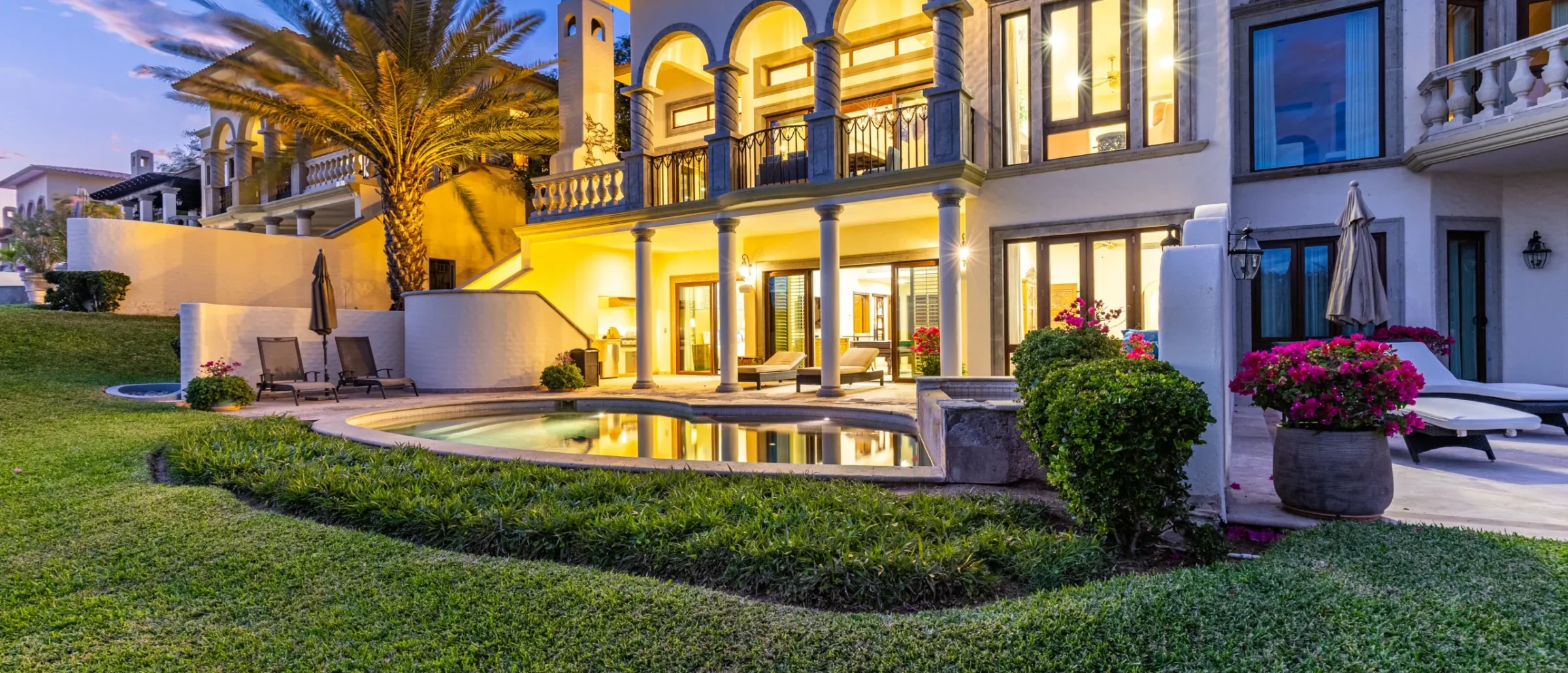 Cream villa at dusk with arched balcony, covered patio, small curved pool, lawn and pink bougainvillea