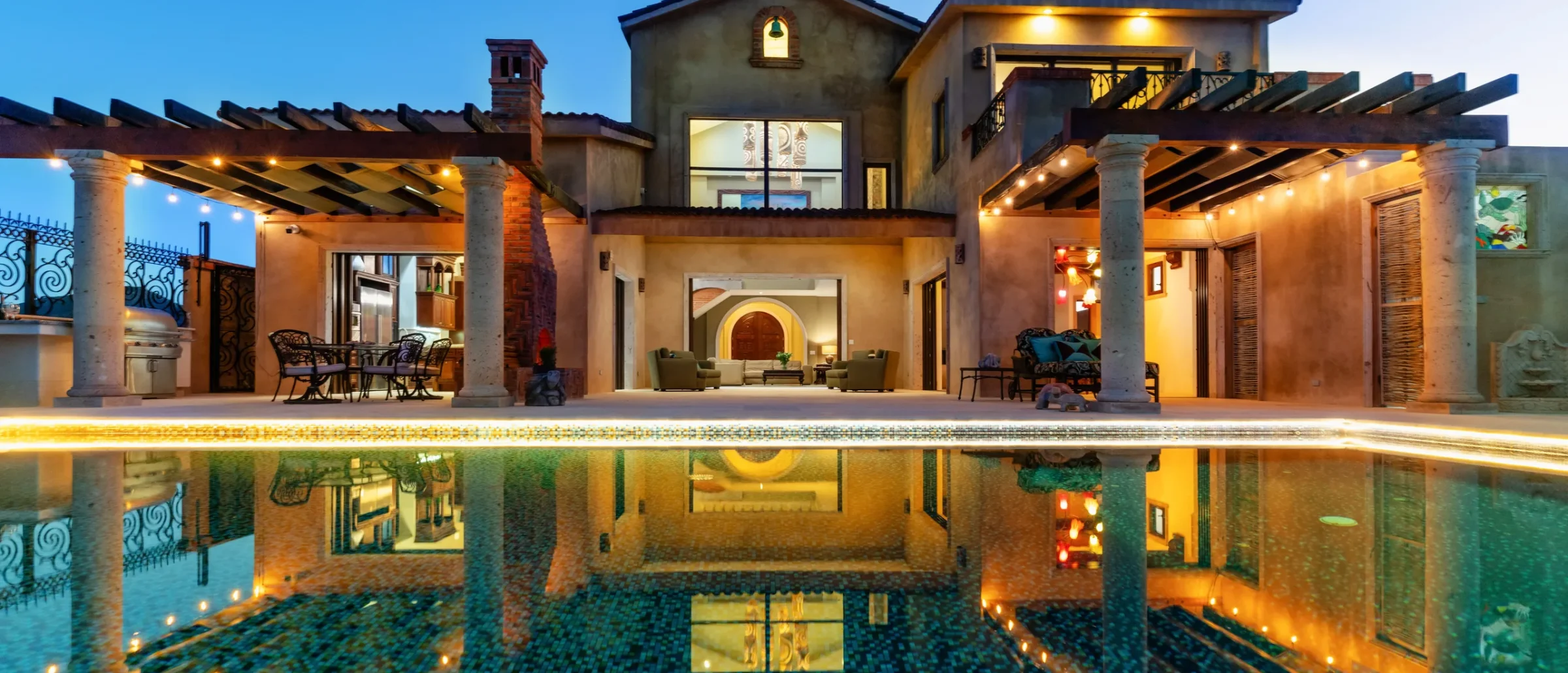 Hacienda-style home at dusk with lit patios under pergolas and a large mosaic pool in front