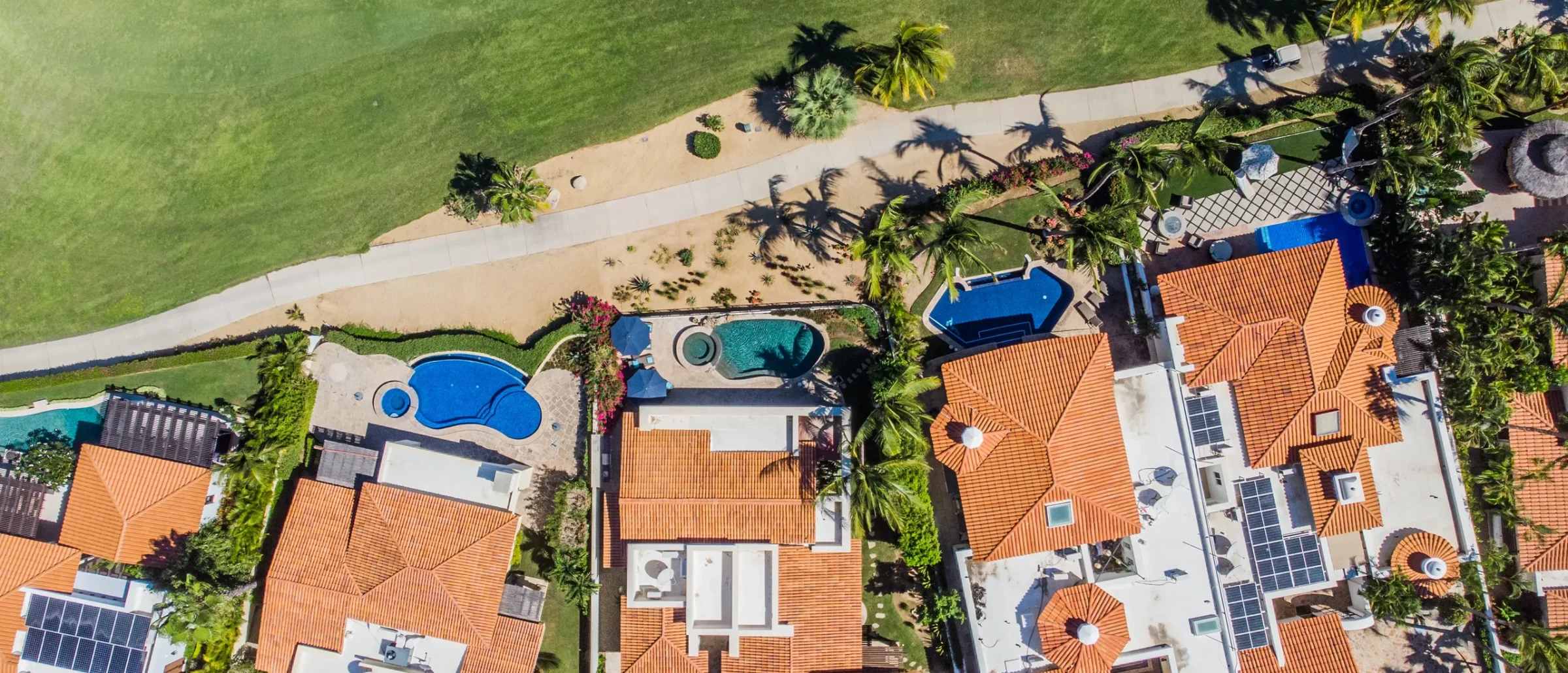 Overhead view of tile-roofed homes with private pools and palms beside a golf course fairway