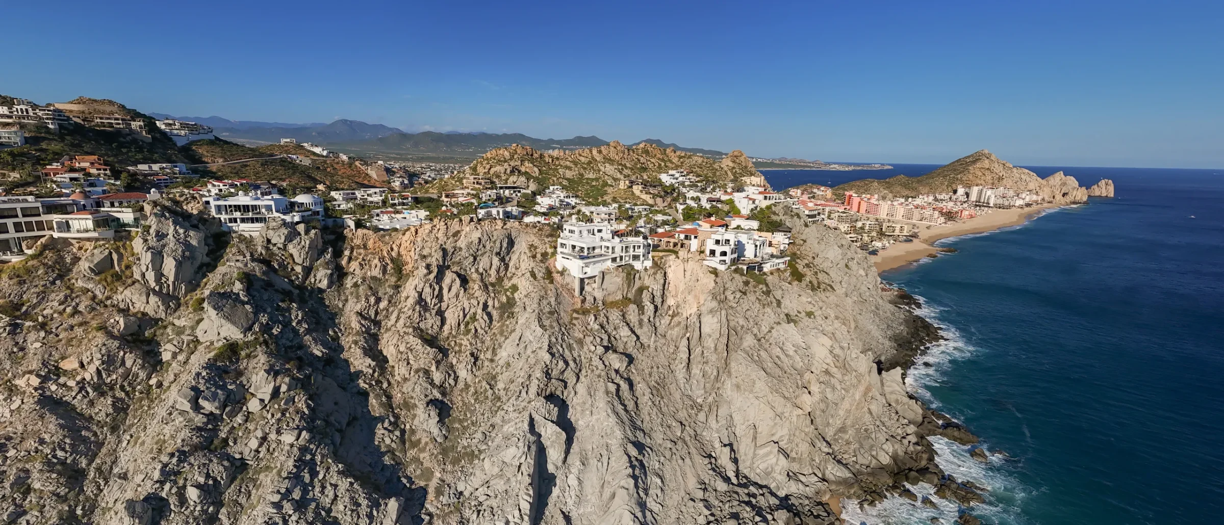 Aerial view of cliffside homes on a granite headland above the ocean, with a hotel-lined beach and rocky point beyond
