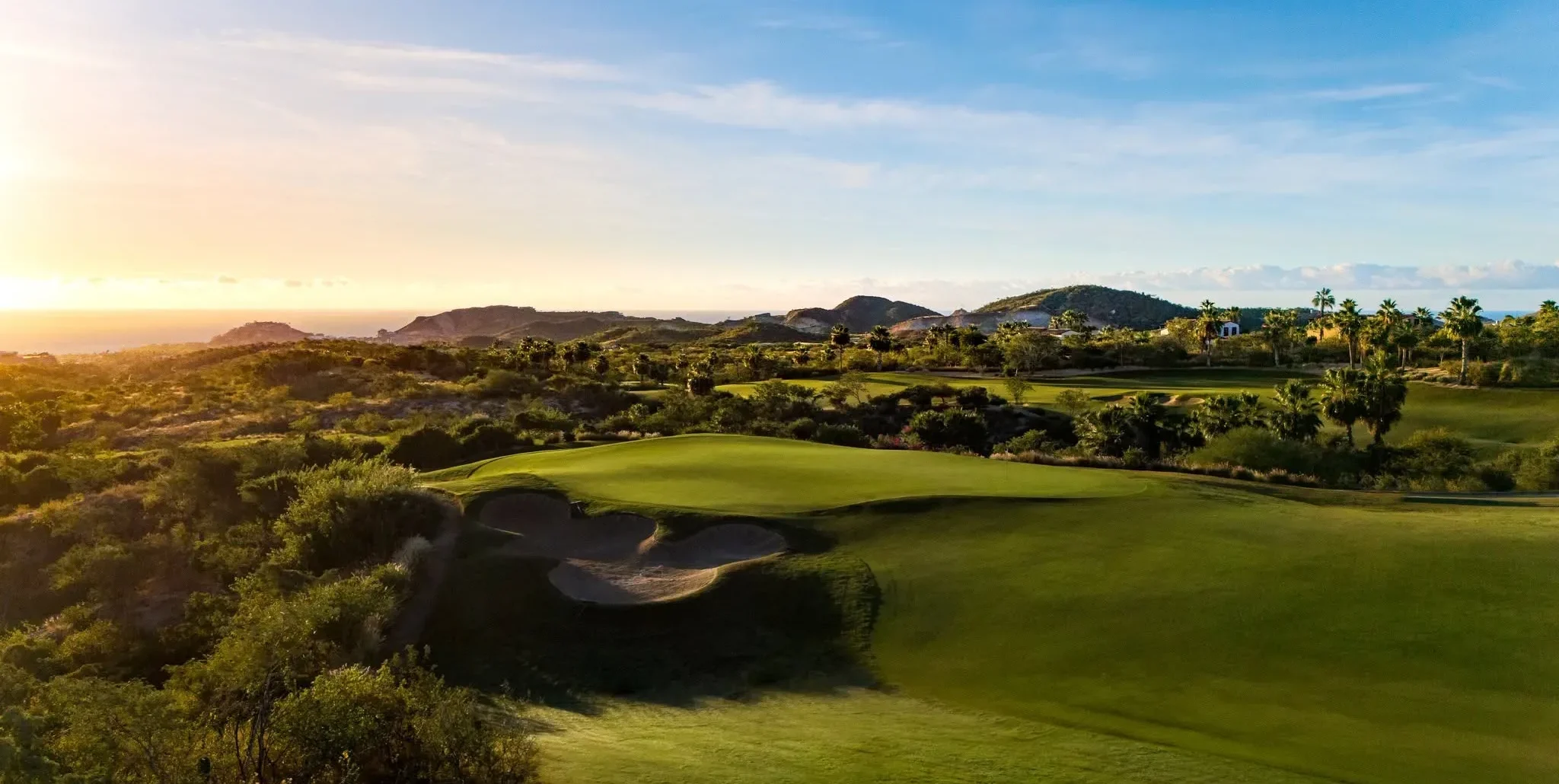 Aerial view of a golf green with sand bunkers, desert hills, palm trees and the ocean at sunset