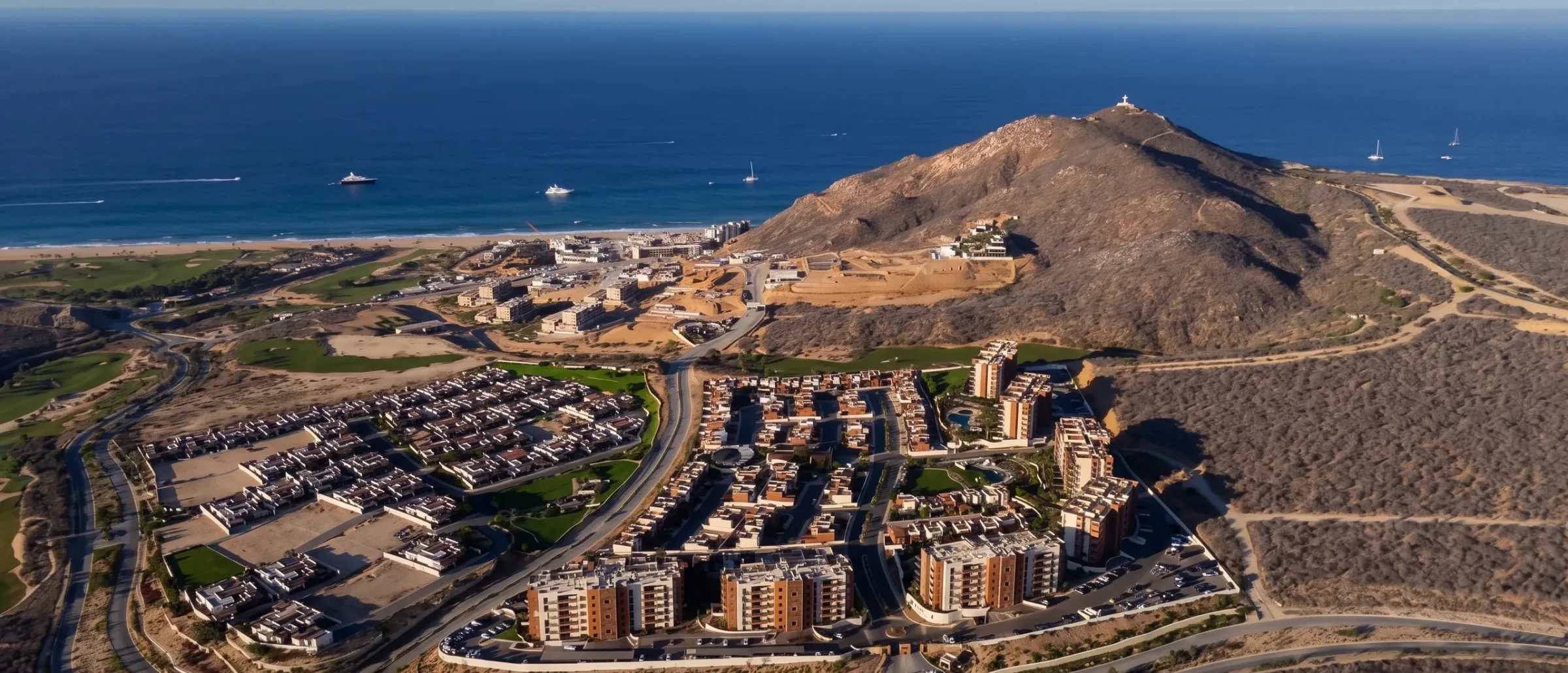 Aerial view of a coastal desert community with condos, townhomes and golf fairways, beside a hill and the ocean