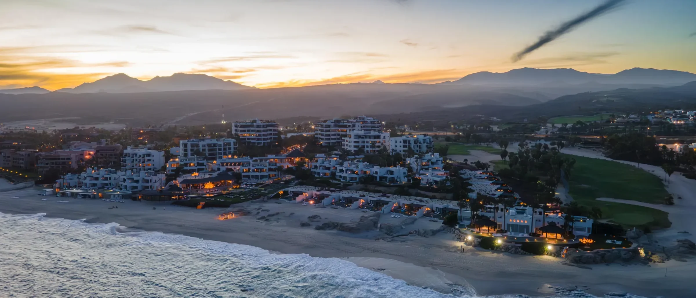 Aerial dusk view of a lit beachfront resort beside a golf course, with ocean waves in front and mountains behind