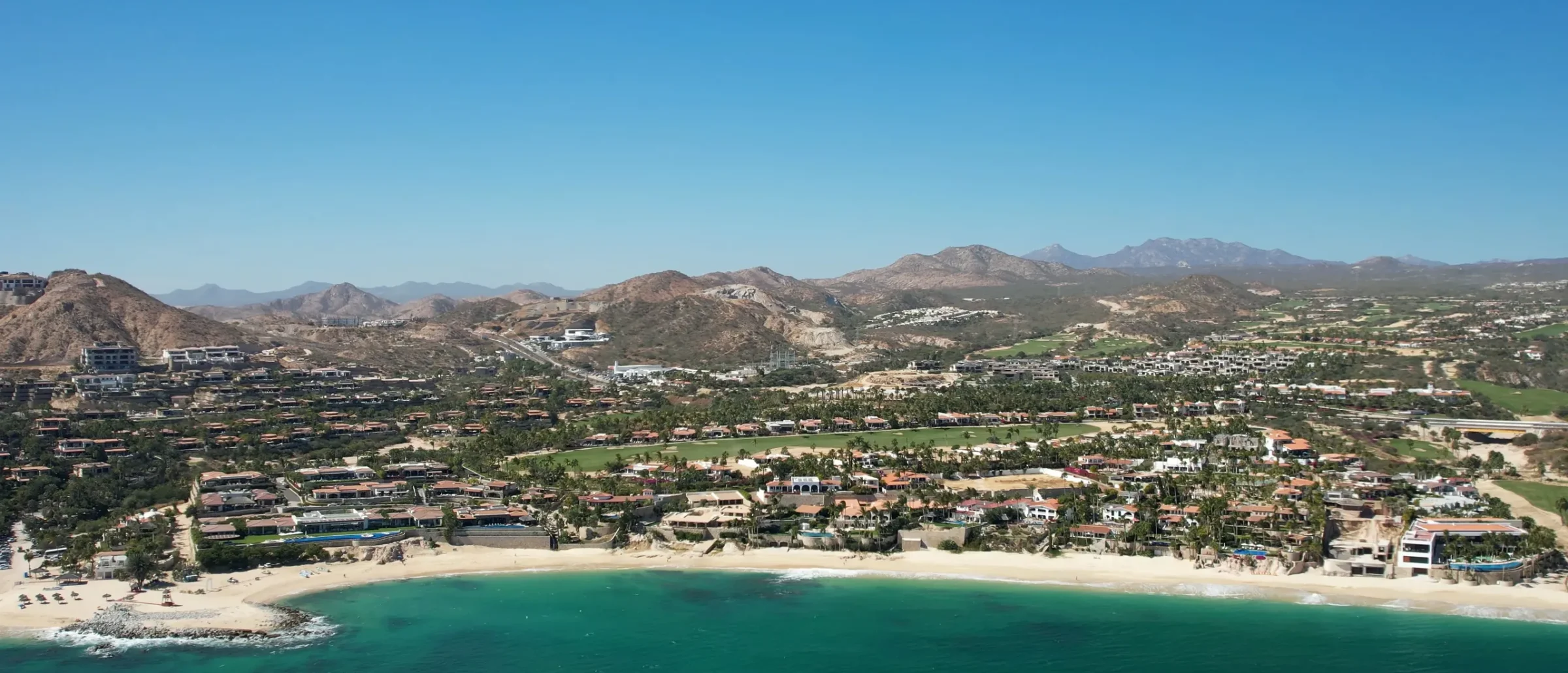 Aerial view of a turquoise bay and sandy beach with homes, palms and a golf course set against dry hills and mountains