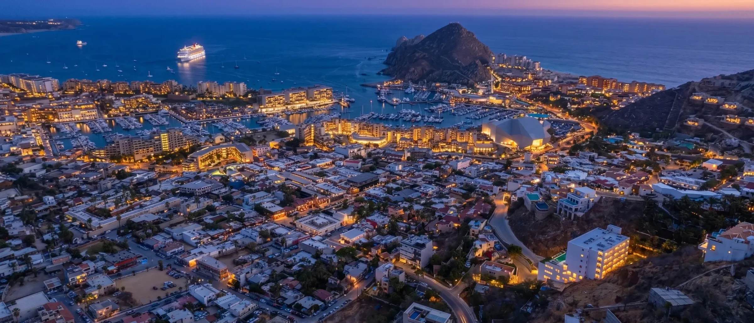 Aerial dusk view of Cabo San Lucas with lit marina, cruise ship in the bay and the rocky Land's End headland