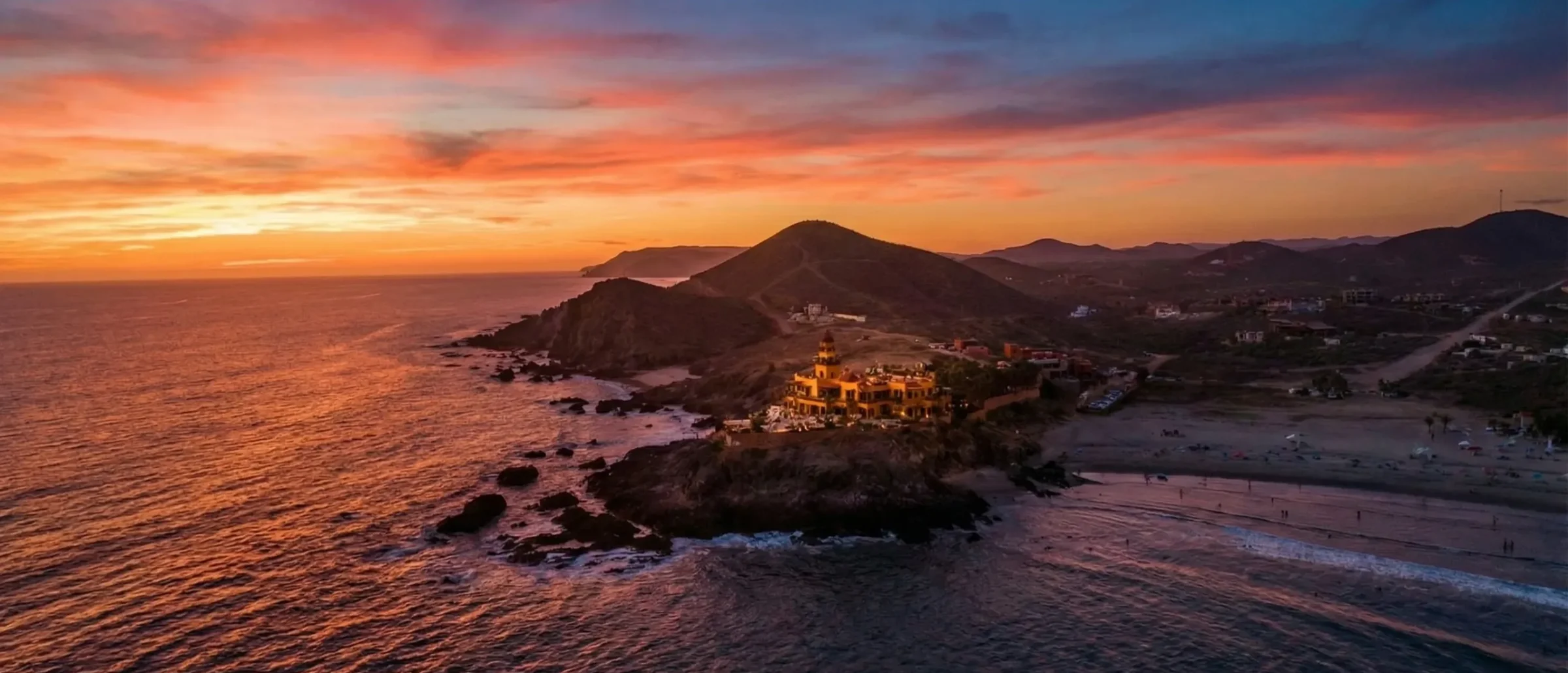 Aerial sunset over a rocky headland and sandy beach on the Pacific coast, with scattered homes and hills inland